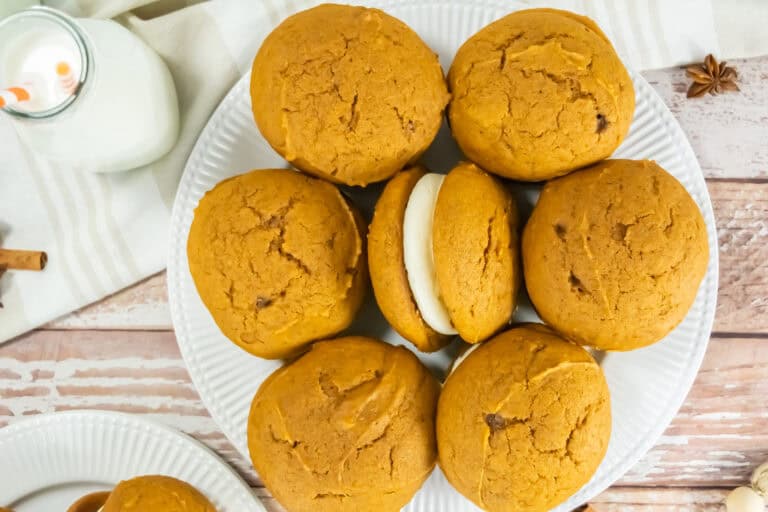 A plate of seven pumpkin whoopie pies with creamy filling is arranged on a wooden table. A glass of milk with straws sits nearby, evoking a cozy autumn feel.