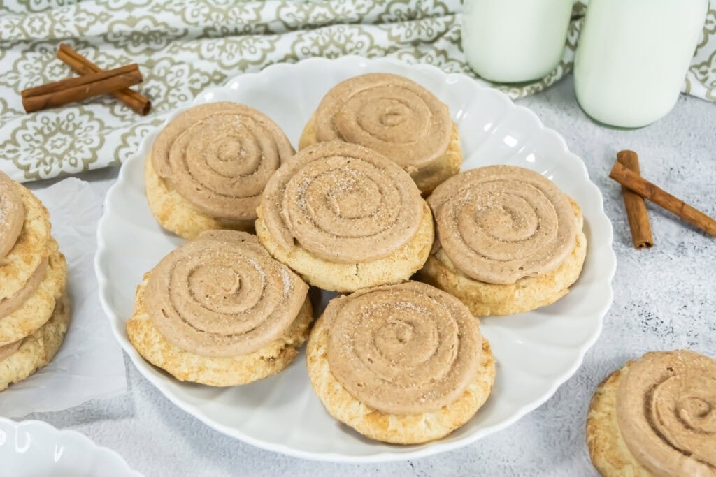 Plate of Crumbl Copycat Churro Cookies topped with swirl frosting, surrounded by cinnamon sticks and two milk bottles, creating a warm, inviting tone.