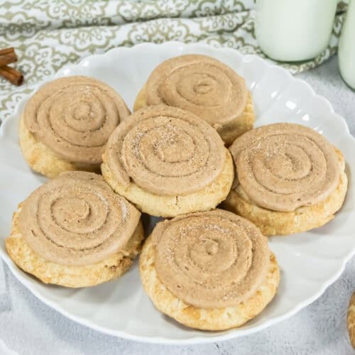 Plate of Crumbl Copycat Churro Cookies topped with swirl frosting, surrounded by cinnamon sticks and two milk bottles, creating a warm, inviting tone.