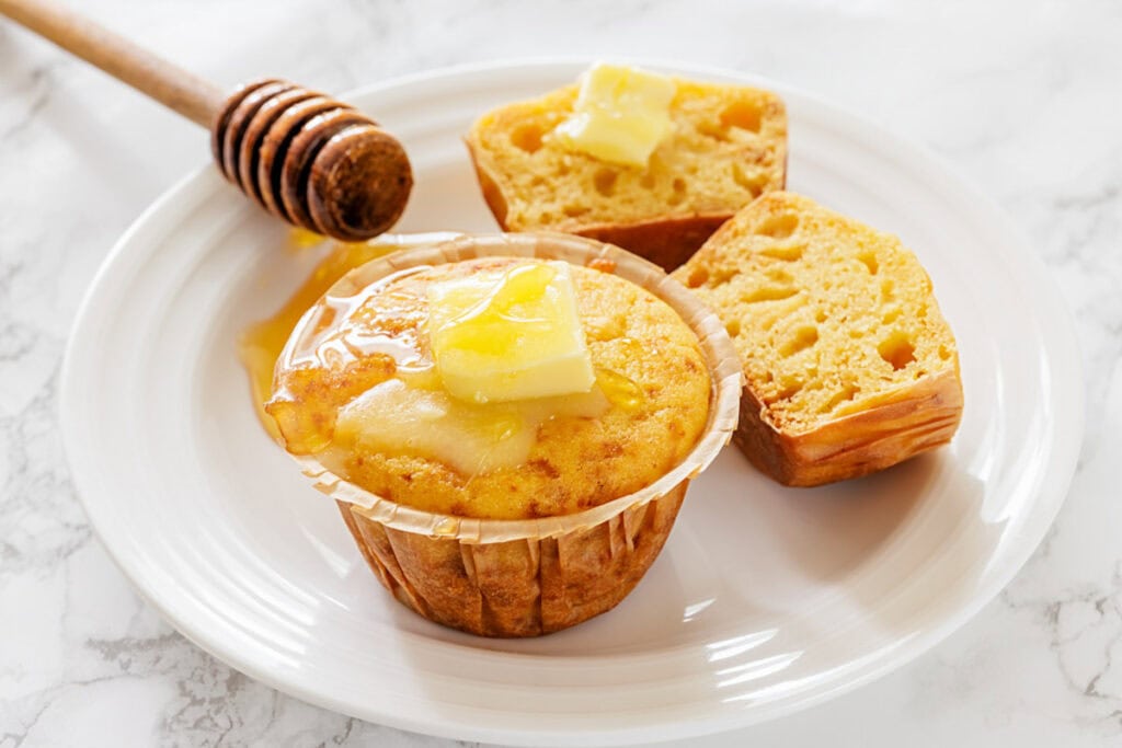 A freshly baked corn muffin on a white plate, topped with melting butter and honey. Nearby, a split muffin half also has butter, exuding warmth and comfort.