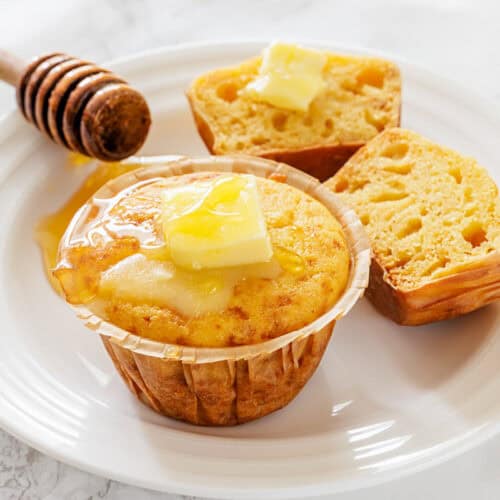 A freshly baked corn muffin on a white plate, topped with melting butter and honey. Nearby, a split muffin half also has butter, exuding warmth and comfort.
