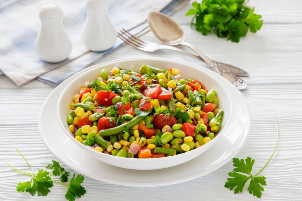 A colorful vegetable salad with tomatoes, corn, edamame, and parsley in a white bowl on a wooden table. Behind are utensils, a napkin, and herbs.