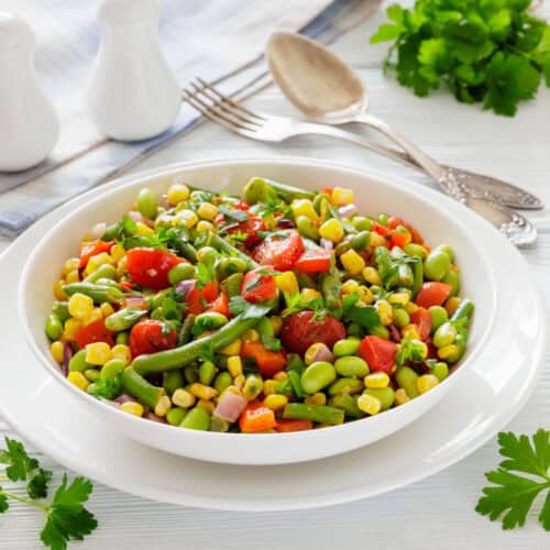 A colorful vegetable salad with tomatoes, corn, edamame, and parsley in a white bowl on a wooden table. Behind are utensils, a napkin, and herbs.