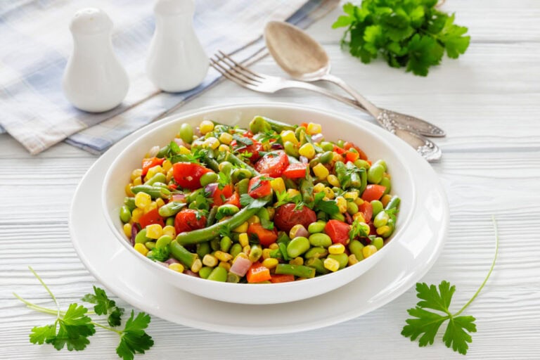 A colorful vegetable salad with tomatoes, corn, edamame, and parsley in a white bowl on a wooden table. Behind are utensils, a napkin, and herbs.