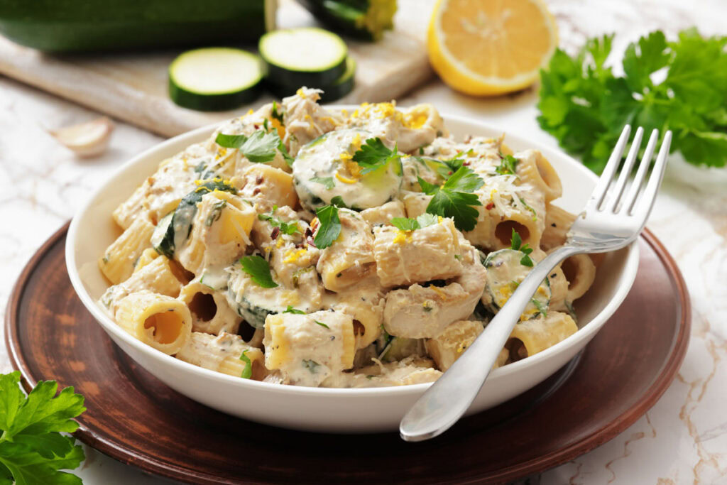 A creamy pasta dish with zucchini, parsley, and lemon zest in a white bowl on a wooden plate. A fork rests on the bowl's rim, creating a fresh, appetizing look.