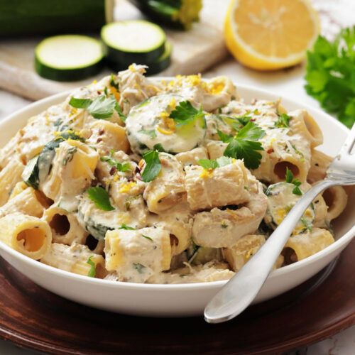 A creamy pasta dish with zucchini, parsley, and lemon zest in a white bowl on a wooden plate. A fork rests on the bowl's rim, creating a fresh, appetizing look.