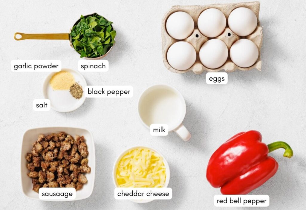 Ingredients for a recipe are neatly arranged on a white surface, including spinach, eggs, garlic powder, pepper, salt, milk, sausage, cheddar cheese, and a red bell pepper.