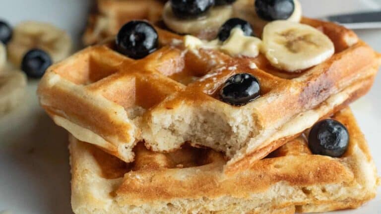 Two stacked waffles with a bite taken out, topped with banana slices, blueberries, and a pat of butter.