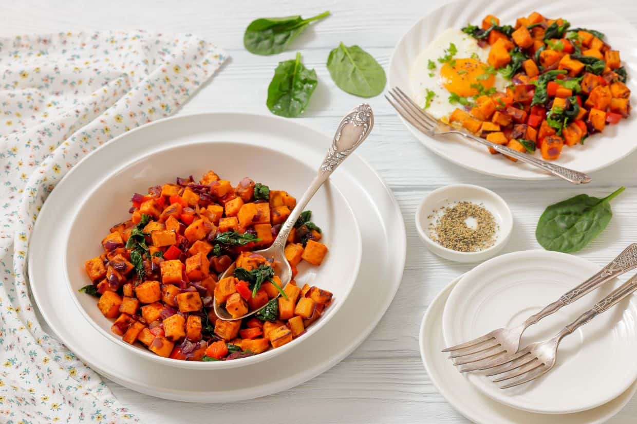 A bowl and plates of sweet potato hash with sautéed spinach and onions, served on a white table with forks, a napkin, and a small bowl of pepper.