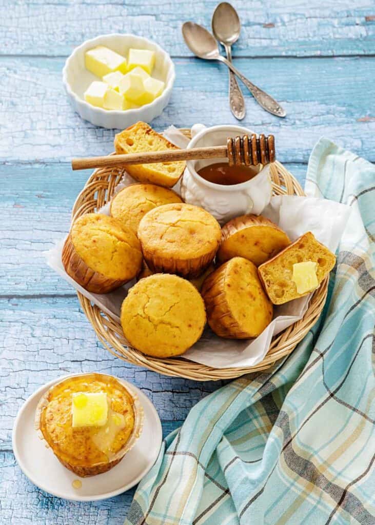 A wicker basket with golden muffins is topped with butter and honey. A dish with butter cubes and two spoons is placed alongside on a rustic blue table.