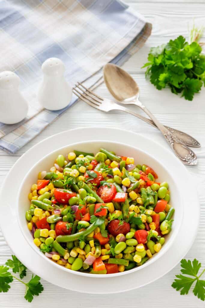 A vibrant salad with cherry tomatoes, corn, edamame, and bell peppers garnished with parsley. Nearby are cutlery, a napkin, and salt shakers.