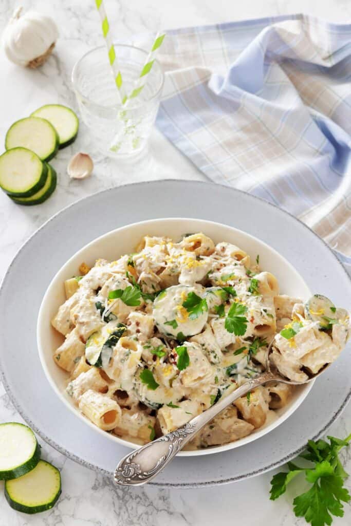 A bowl of creamy pasta with zucchini and parsley, garnished with lemon zest. Surrounded by sliced zucchini, garlic, a drink, and a checkered cloth.