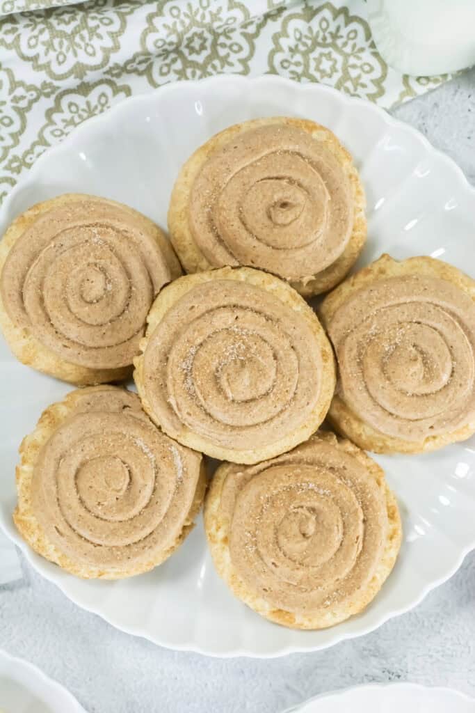 Plate of six cinnamon sugar cookies with swirled frosting, arranged in a circle on a white, scalloped dish. Background features an elegant patterned cloth.