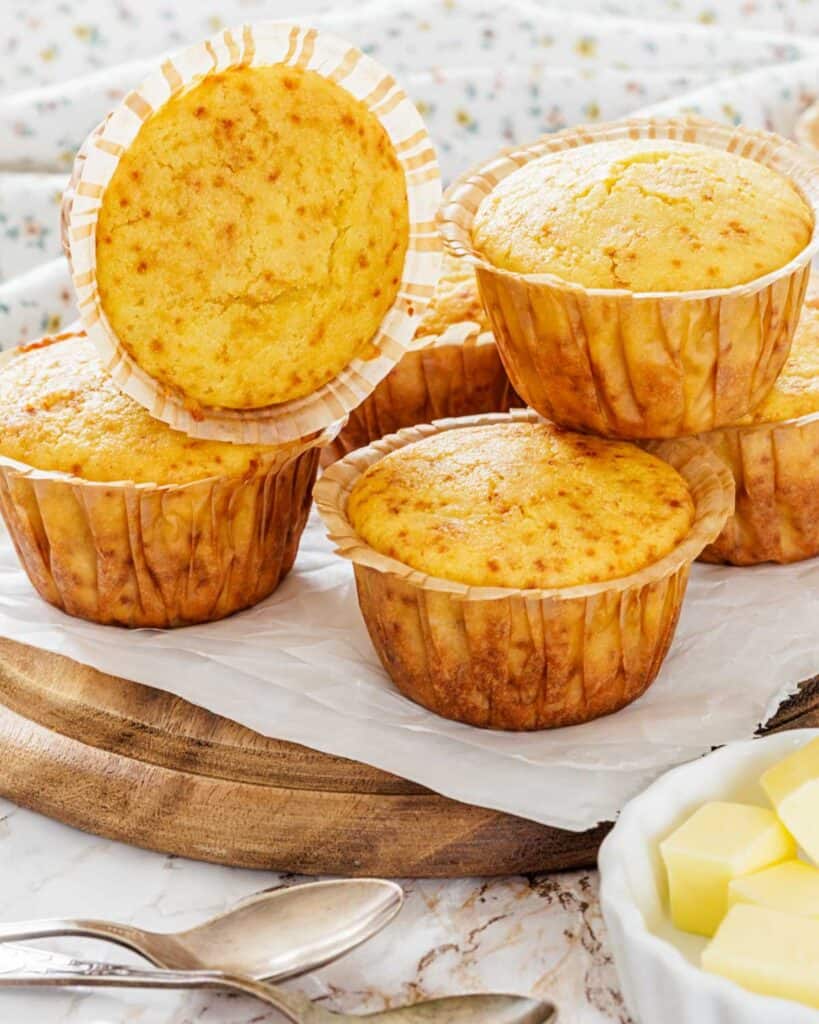 Golden corn muffins are stacked on a wooden board lined with parchment paper. A bowl of butter cubes and silver spoons are placed beside them.
