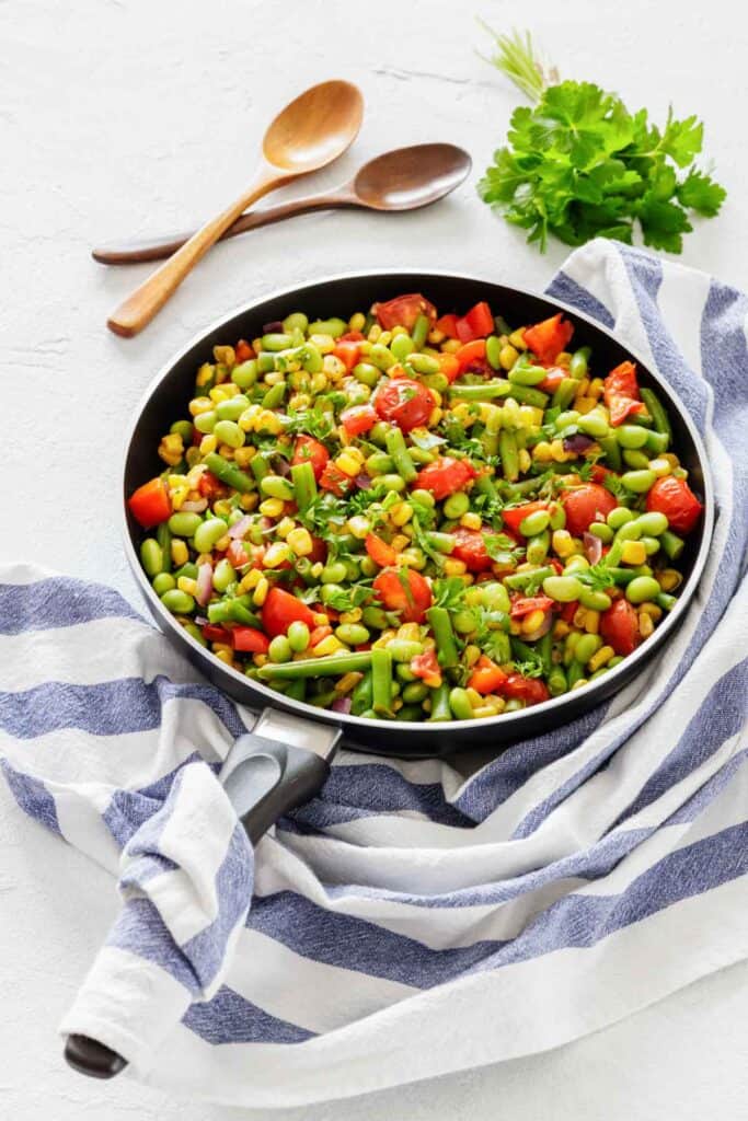 A colorful skillet of sautéed vegetables, including edamame, corn, and cherry tomatoes, rests on a striped cloth with wooden utensils and fresh parsley nearby.