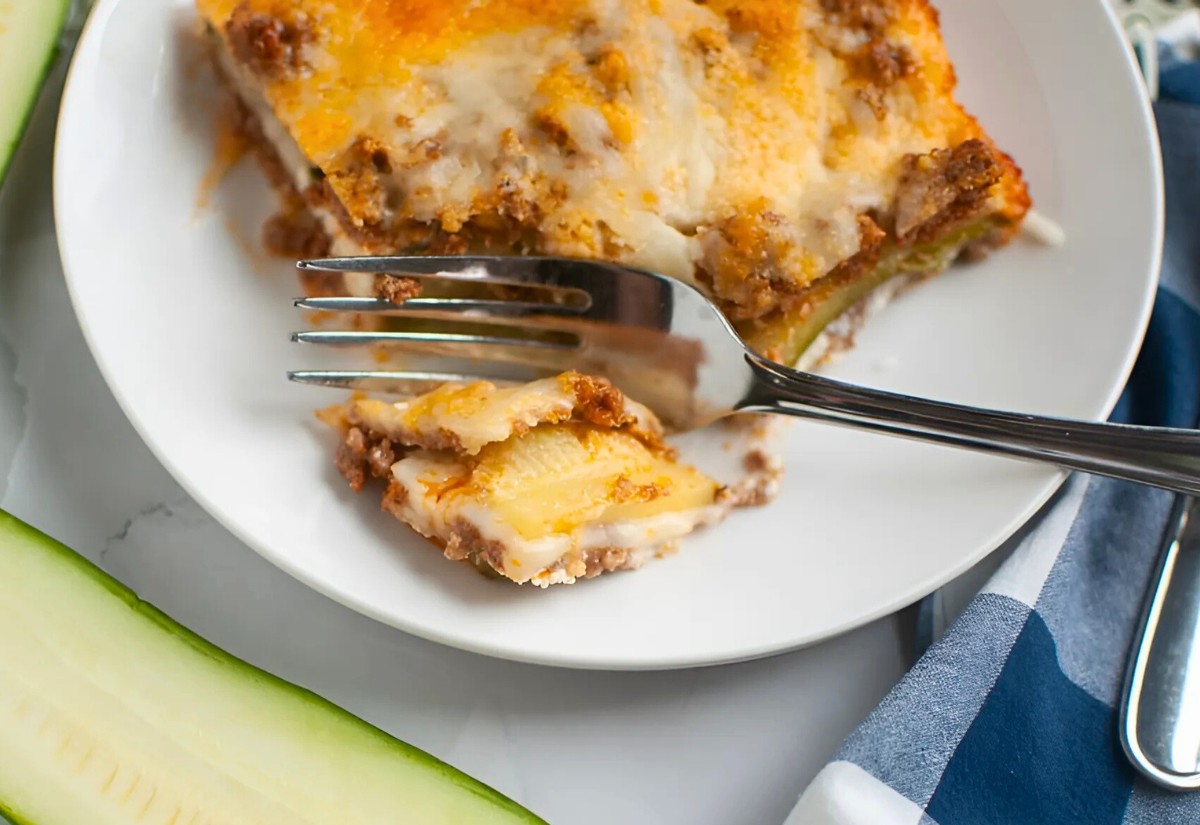 A slice of zucchini lasagna on a white plate with a fork holding a piece, next to fresh zucchini slices and a blue checkered napkin.