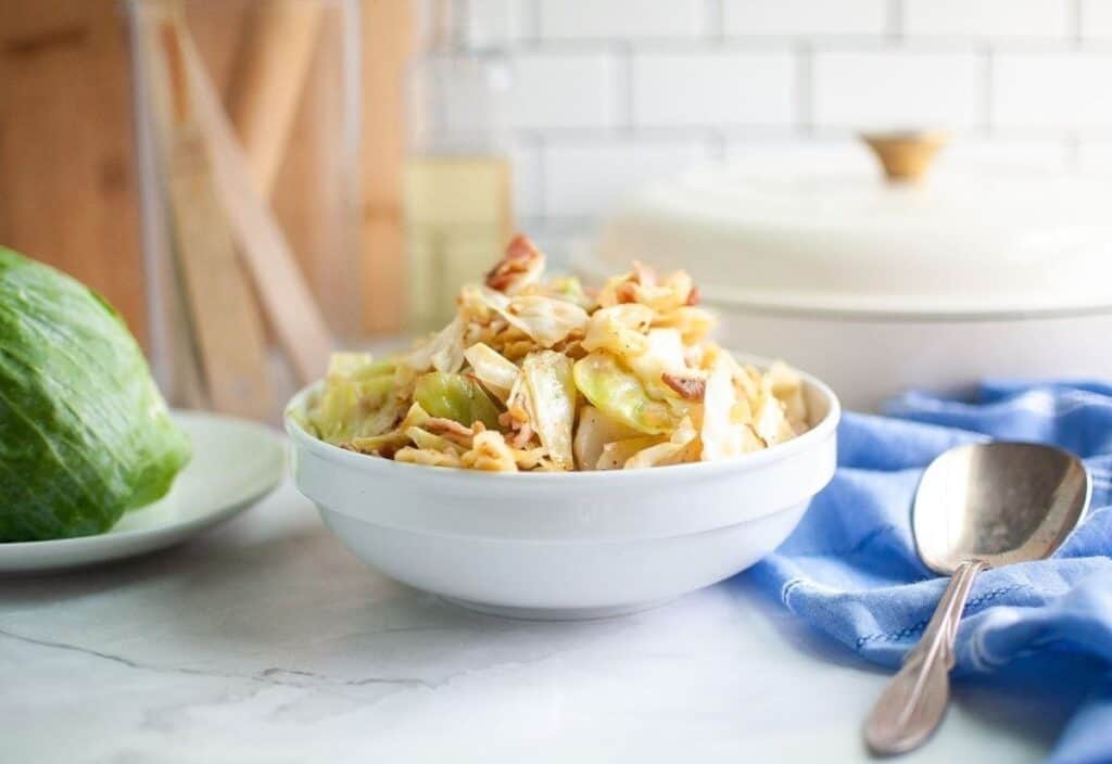 A bowl of sautéed cabbage and bacon on a white counter with a spoon on a blue cloth, fresh cabbage nearby, creating a cozy kitchen setting.
