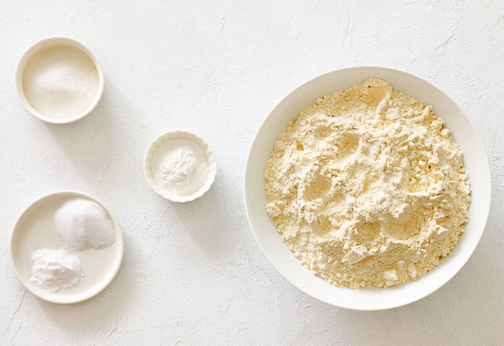 A bowl of flour with sugar, salt, and baking powder arranged beside it.
