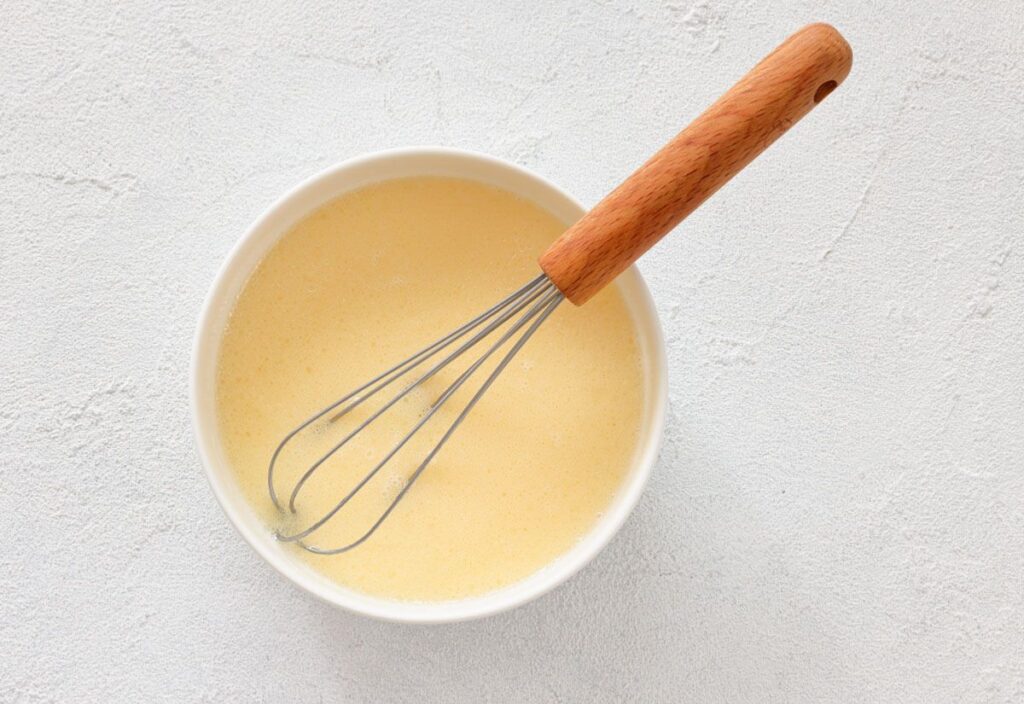 A bowl of a liquid mixture with a whisk resting inside.