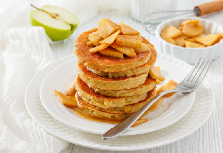 A stack of golden brown pancakes topped with cinnamon-spiced apple slices on a white plate. A fork rests beside them. Background includes a green apple half and a bowl of apples, creating a cozy, inviting breakfast scene.