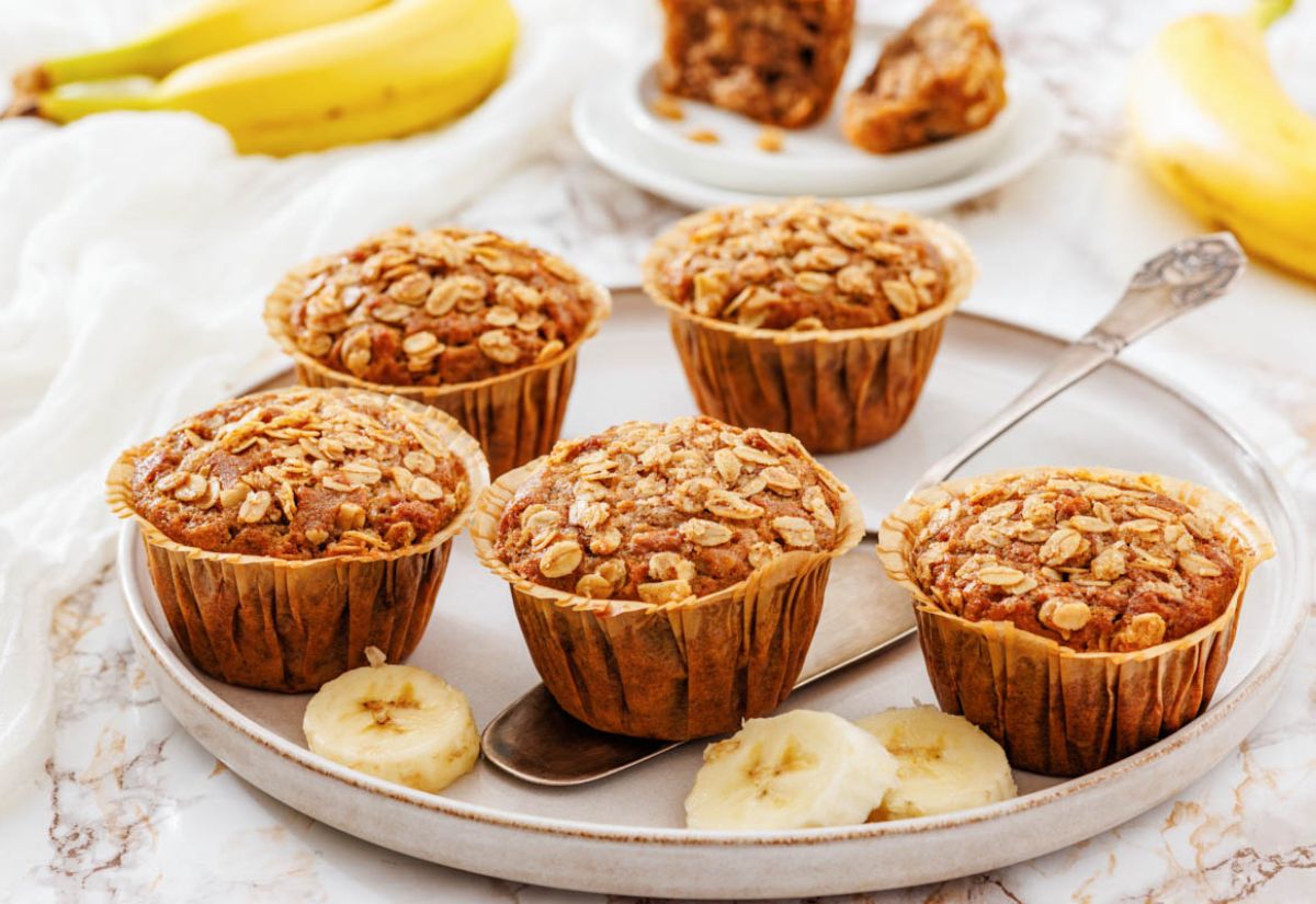 Five banana muffins with oats on top sit on a round tray with sliced bananas. A spoon and whole bananas are in view. Warm, inviting tone.