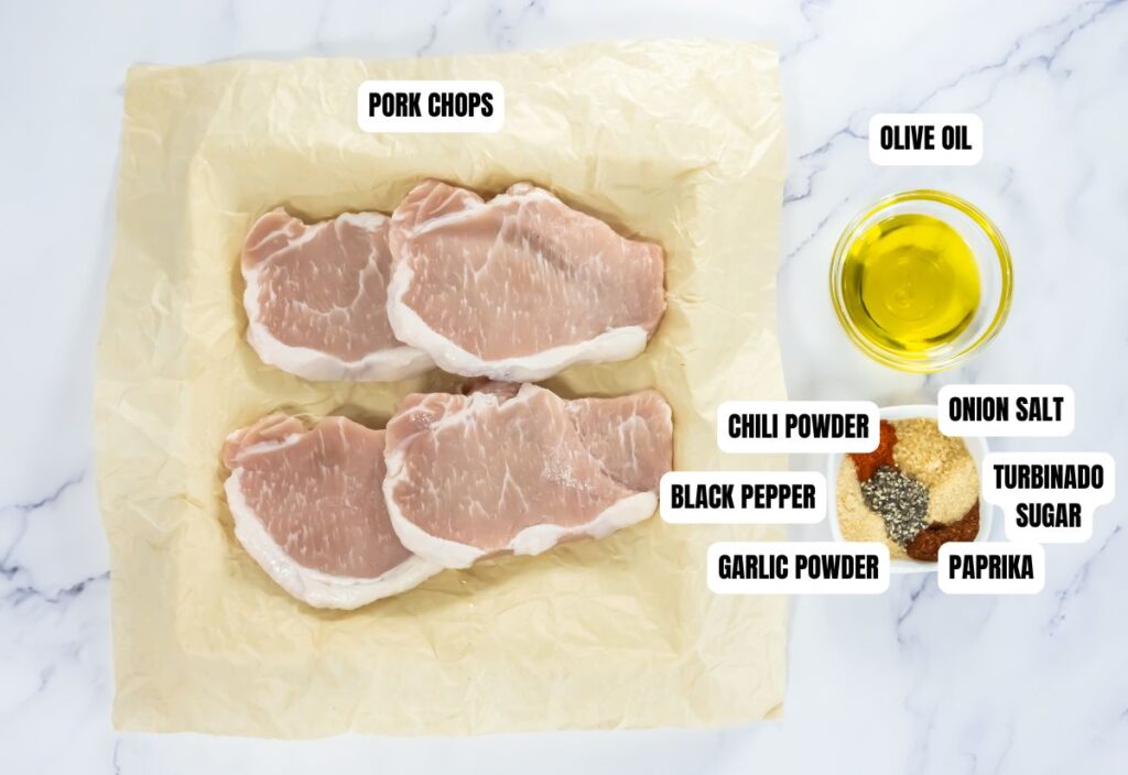 Raw pork chops on parchment with labeled spices: chili powder, black pepper, garlic powder, onion salt, turbinado sugar, paprika, and olive oil.