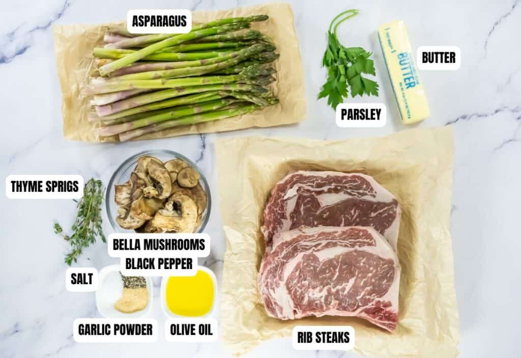 A flat lay of ingredients on a marble surface includes rib steaks, asparagus, mushrooms, thyme, parsley, butter, garlic powder, olive oil, salt, and black pepper.