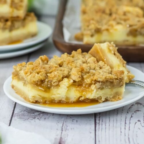 Square apple cheesecake bar with a crumbly top, creamy layer, and caramel drizzle on a white plate. Other bars and caramel sauce visible in the background.
