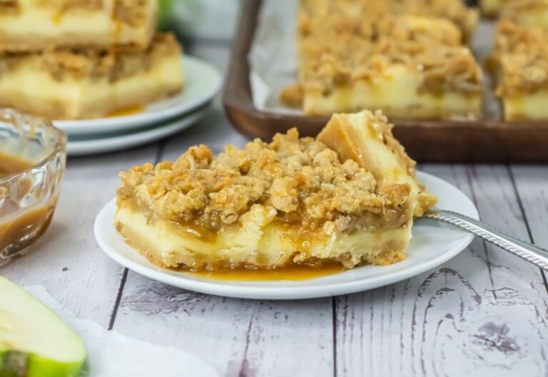 Square apple crumble bar with a crumbly top, creamy layer, and caramel drizzle on a white plate. Other bars and caramel sauce visible in the background.