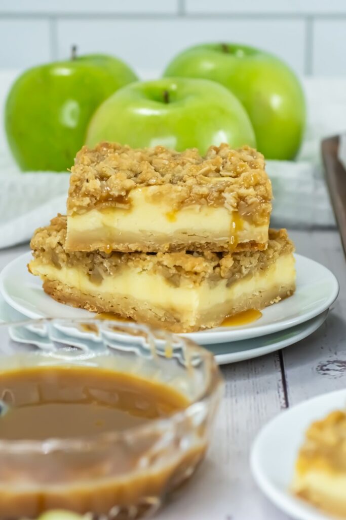 Two caramel apple cheesecake bars stacked on a plate, topped with oat crumble. Fresh green apples and caramel sauce are visible in the background.