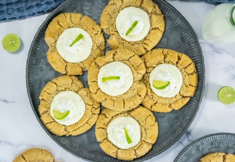A plate of six cookies, each topped with a dollop of white cream and a lime slice, rests on a marble surface, accompanied by halved limes and a bottle of milk.