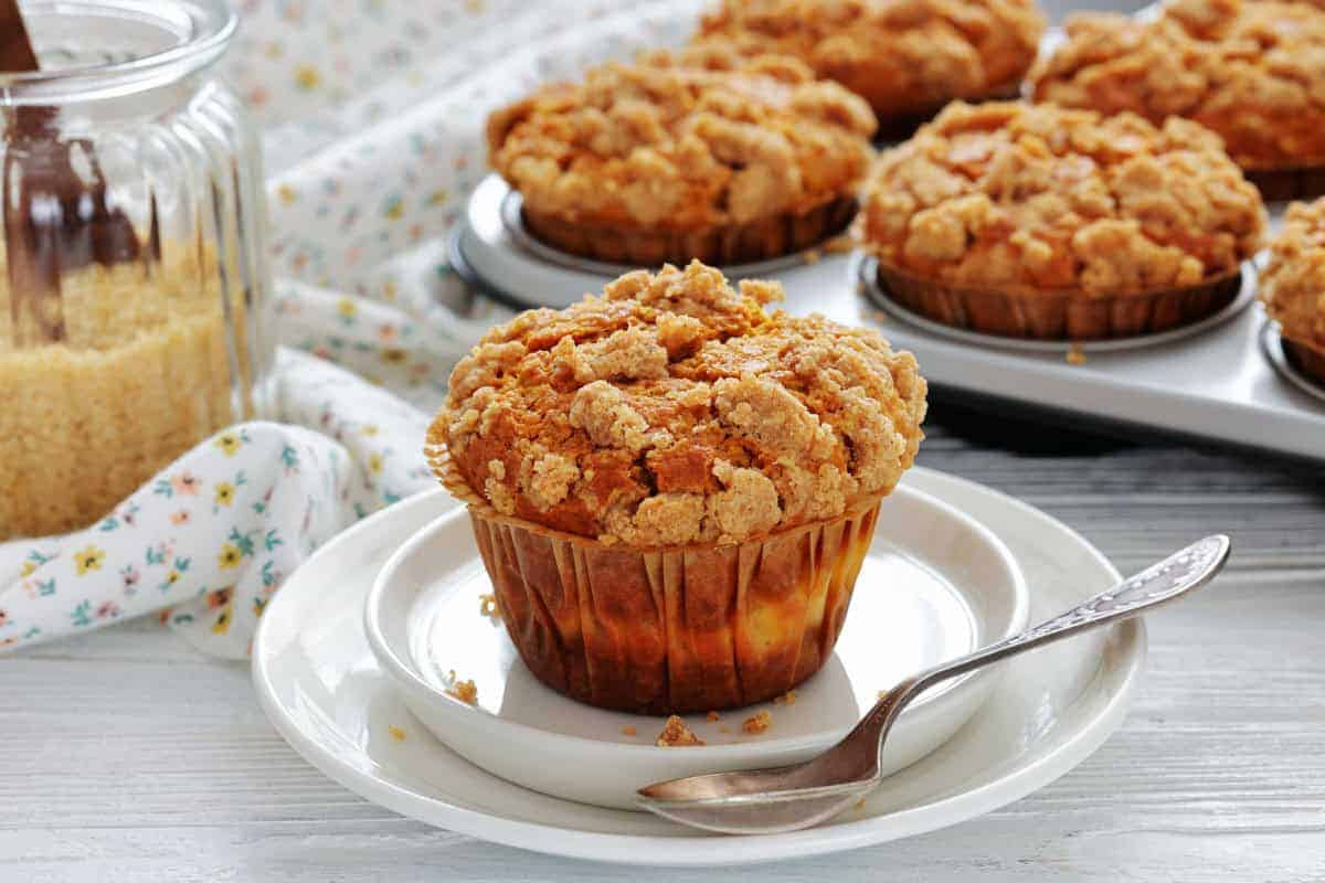 A crumb-topped muffin sits on a white plate with a spoon; more muffins are in a baking tray in the background, next to a jar of brown sugar.