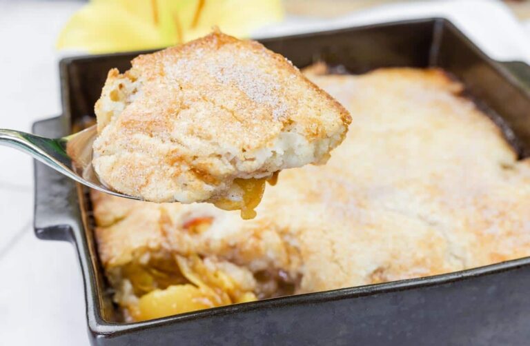A close-up of a spoon holding a serving of peach cobbler above a baking dish filled with the dessert.
