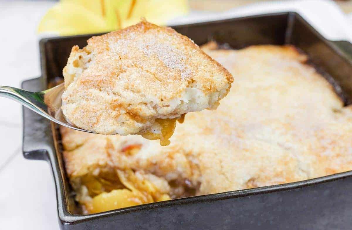 A close-up of a spoon holding a serving of peach cobbler above a baking dish filled with the dessert.