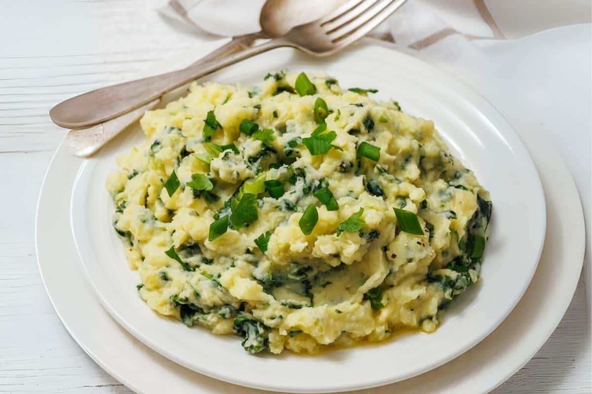 A white plate with colcannon and kale with green herbs is garnished with chopped scallions