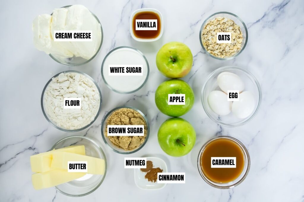 Top-down view of baking ingredients on marble: cream cheese, flour, butter, vanilla, sugar, brown sugar, oats, cinnamon, nutmeg, green apples, eggs, caramel.