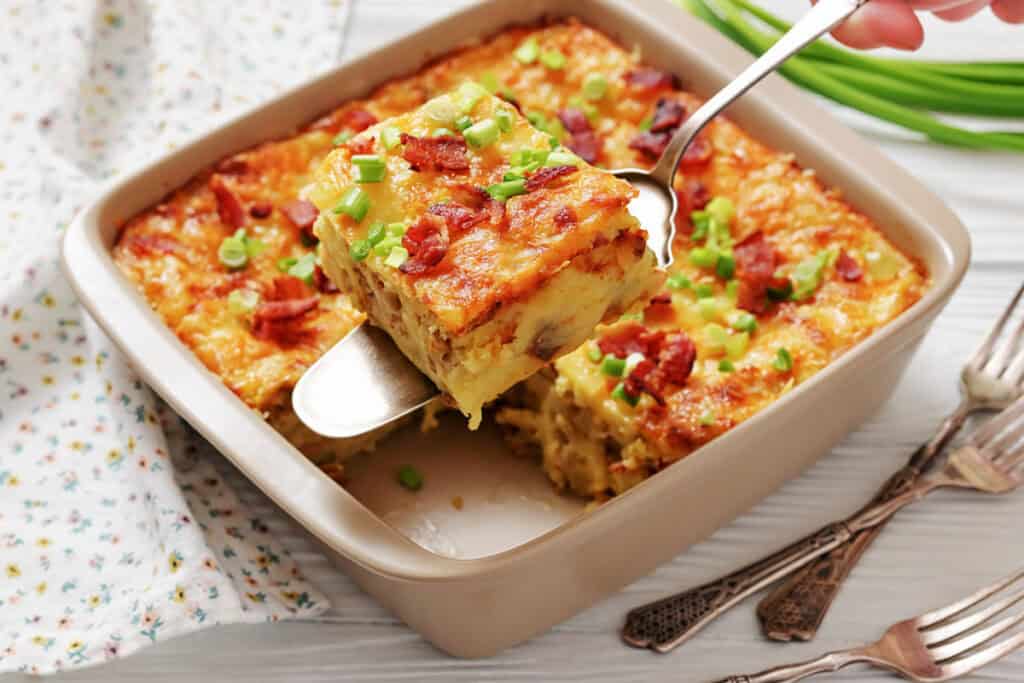 A baked casserole topped with chopped green onions and bacon pieces, with one square being served from the dish using a spatula.