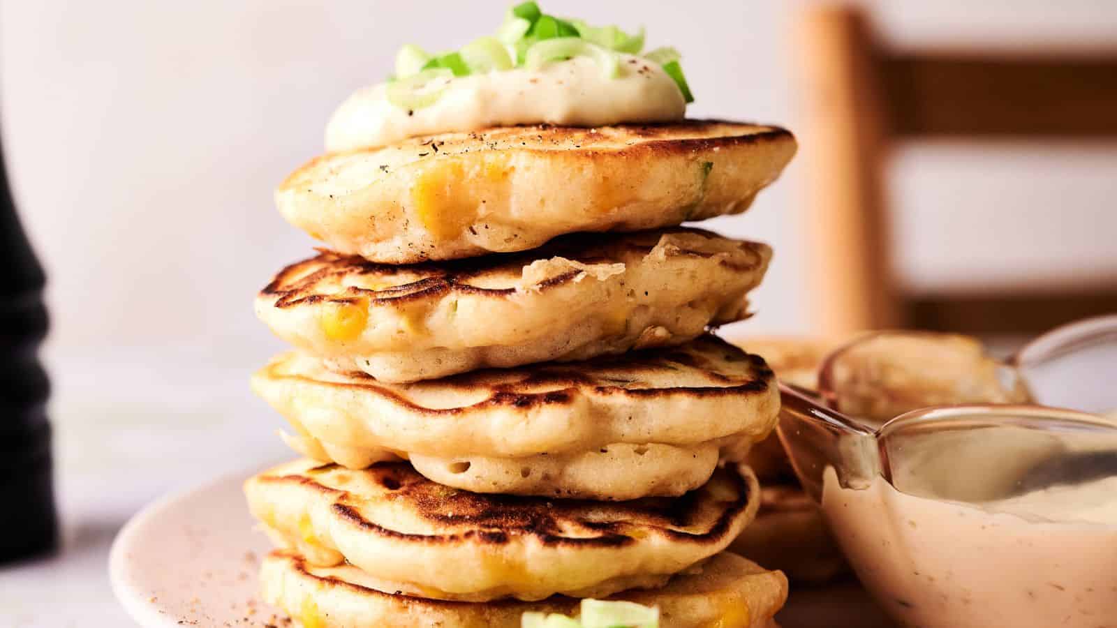 A stack of corn fritters topped with a dollop of cream and chopped green onions, served on a plate with a bowl of dipping sauce nearby.