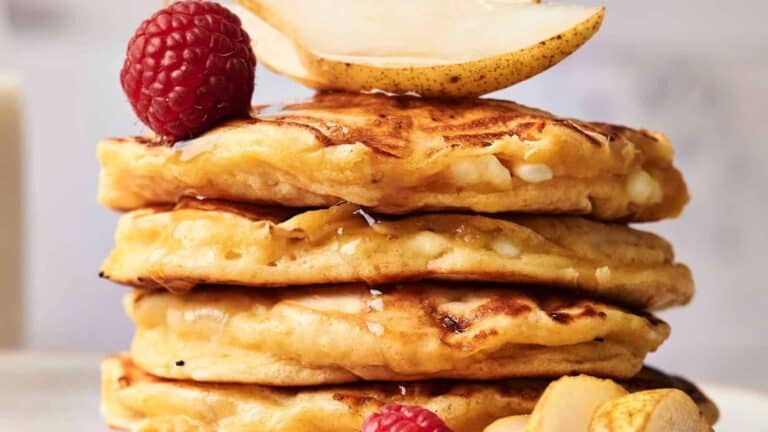 A stack of four fluffy pancakes topped with fresh raspberries, pear slices, and a drizzle of syrup.