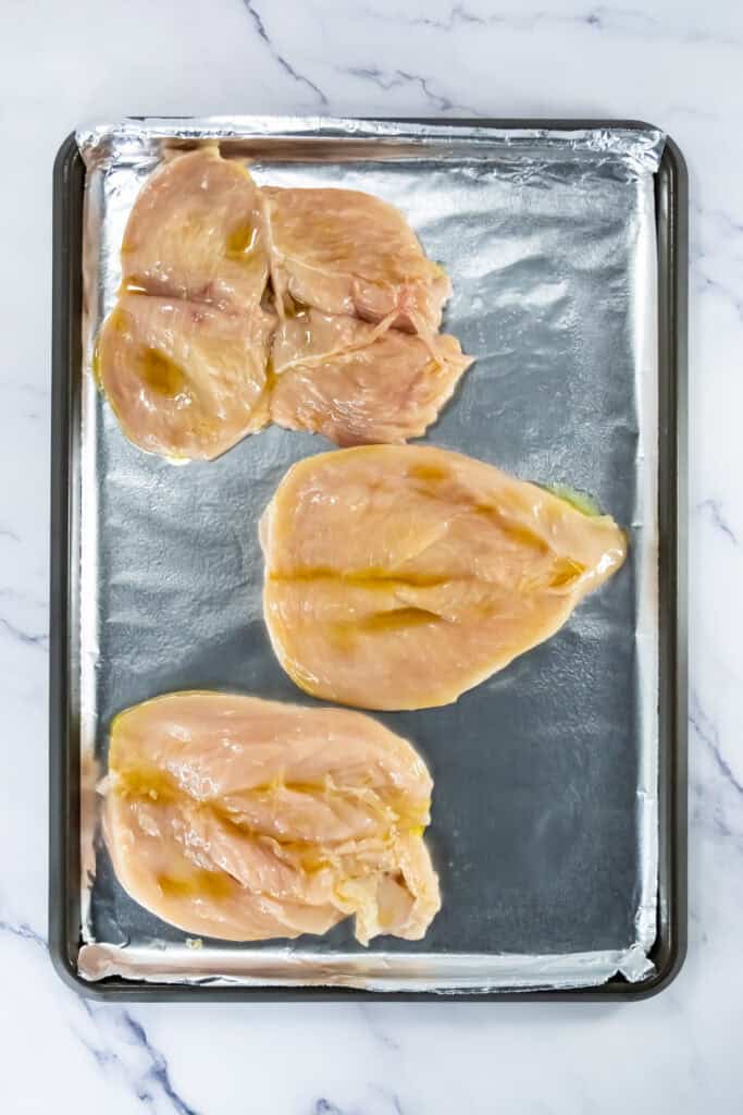 Butterflied raw chicken breasts on a foil-lined baking sheet brushed with olive oil.