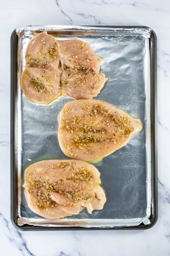 Butterflied raw chicken breasts on a foil-lined baking sheet seasoned with a garlic, cayenne pepper, black pepper, and salt mixture.