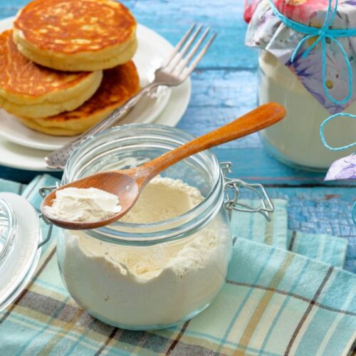 A glass jar with a spoon of white powder, likely flour, on a plaid cloth. Nearby, a plate of pancakes and two jars with floral fabric covers on a blue table.
