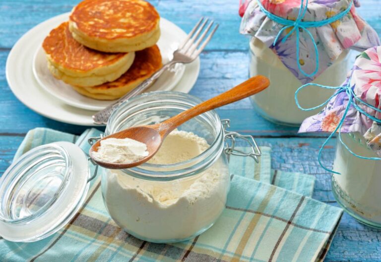 A glass jar with a spoon of white powder, likely flour, on a plaid cloth. Nearby, a plate of pancakes and two jars with floral fabric covers on a blue table.
