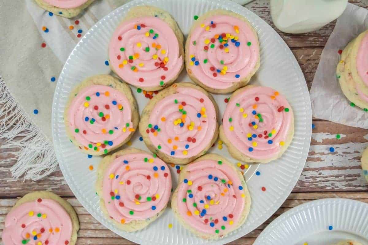 A plate of Copycat Crumbl Confetti Cake Cookies topped with pink frosting and multicolored sprinkles sits on a wooden table.