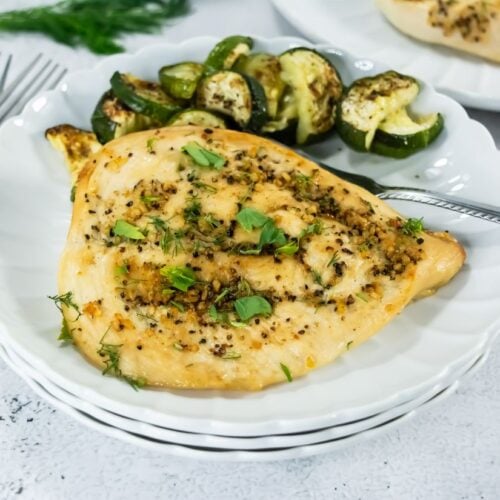 Broiled chicken breast on a white plate, garnished with herbs and spices, served with roasted zucchini. Utensils on the side, set on a white table.
