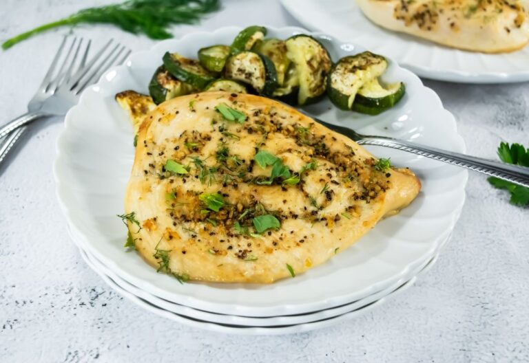 Broiled chicken breast on a white plate, garnished with herbs and spices, served with roasted zucchini. Utensils on the side, set on a white table.