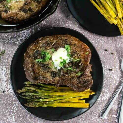 Grilled steak topped with butter and herbs on a black plate with roasted asparagus, set on a textured surface. Nearby, a fork and knife rest.