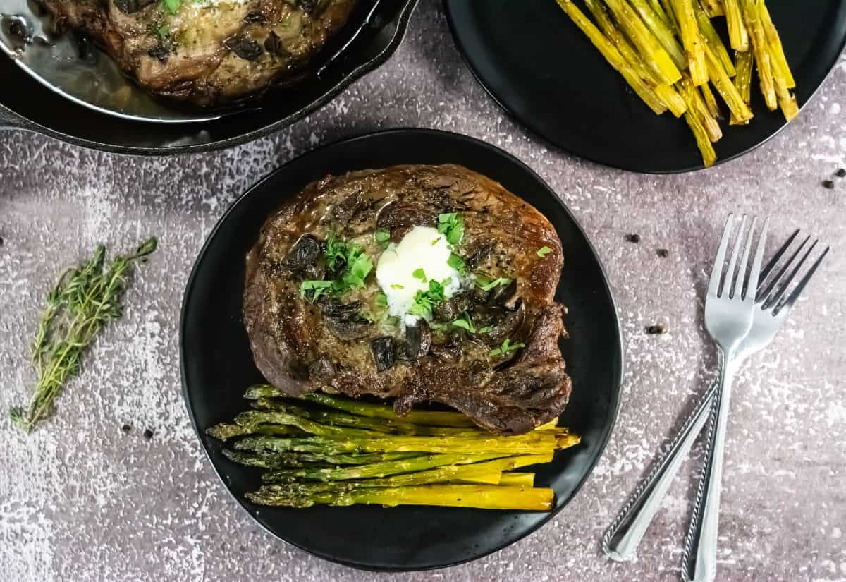 Grilled steak topped with butter and herbs on a black plate with roasted asparagus, set on a textured surface. Nearby, a fork and knife rest.