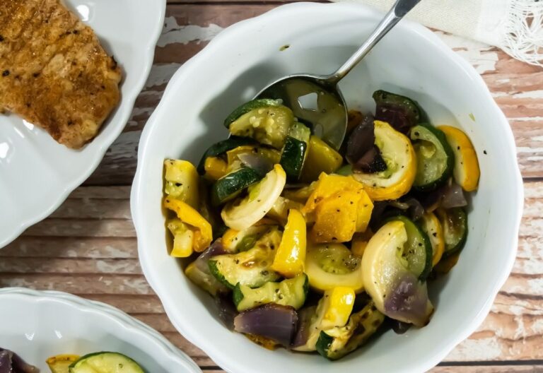 A white bowl filled with colorful roasted vegetables, including zucchini, yellow squash, and red onions, with a spoon. A piece of cooked meat is nearby on a wooden table.