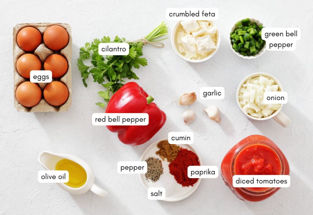 Eggs, cilantro, red and green bell peppers, crumbled feta, garlic, onion, spices, and olive oil arranged on a white surface, suggesting a fresh and vibrant cooking scene.